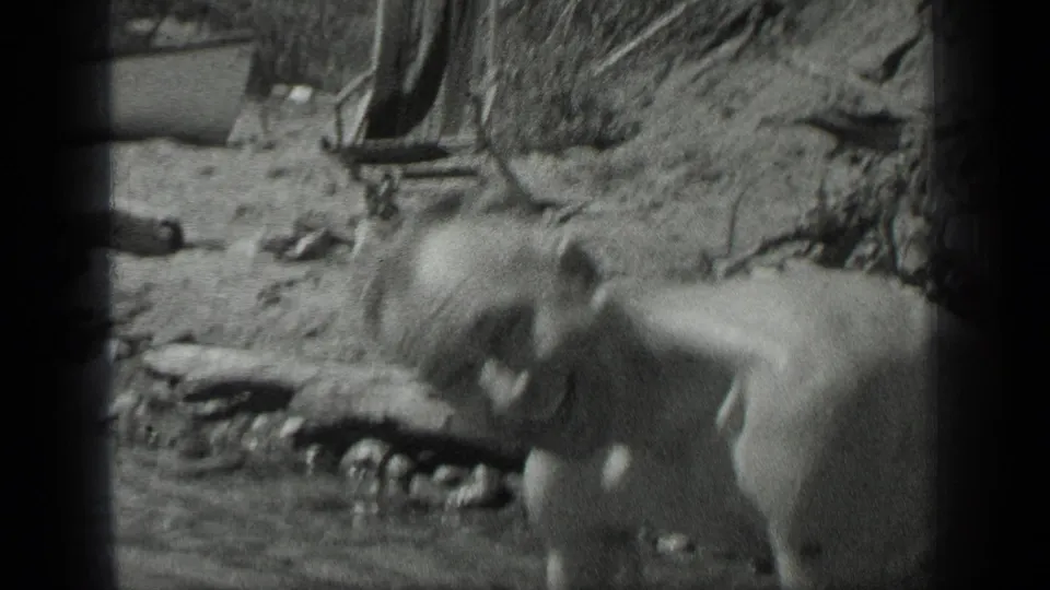 1939 Long Lake, Canada frame showing a man reclining or leaning near the rocky lakeshore, with a wooden chair visible behind him.