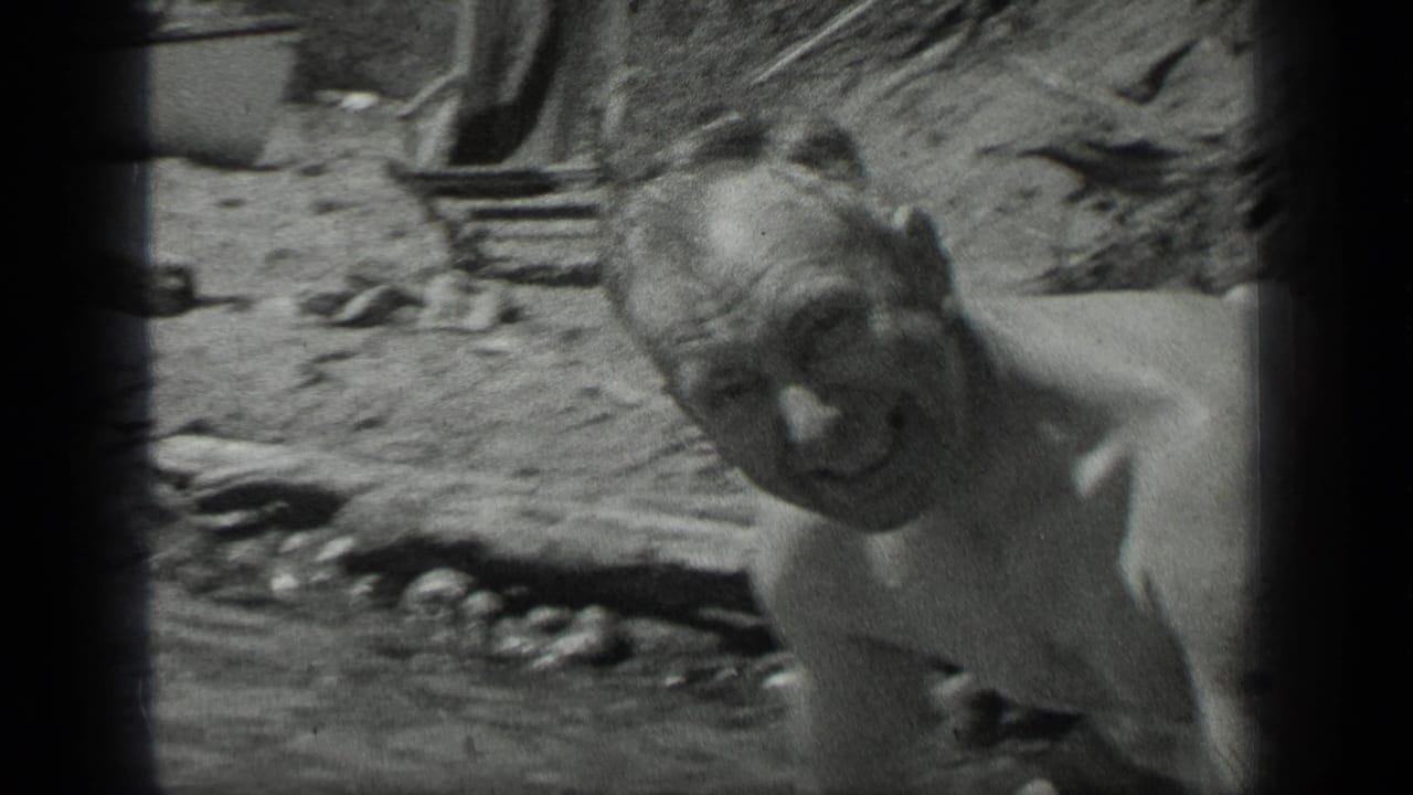 At Long Lake, Canada in 1939, a shirtless adult man smiles toward the camera from shallow water near a rough lakeside shore.