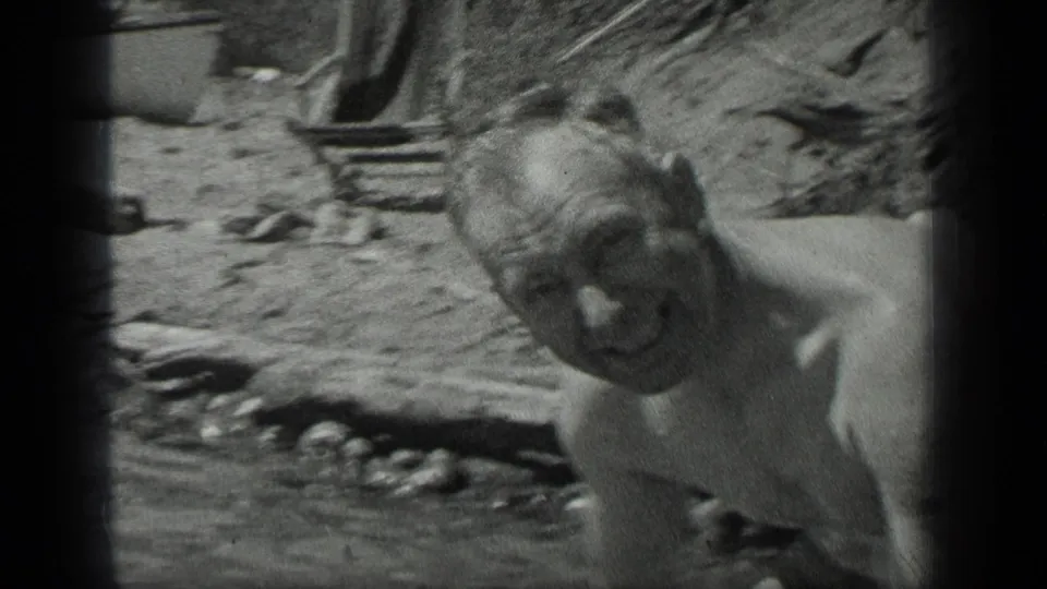 At Long Lake, Canada in 1939, a shirtless adult man smiles toward the camera from shallow water near a rough lakeside shore.