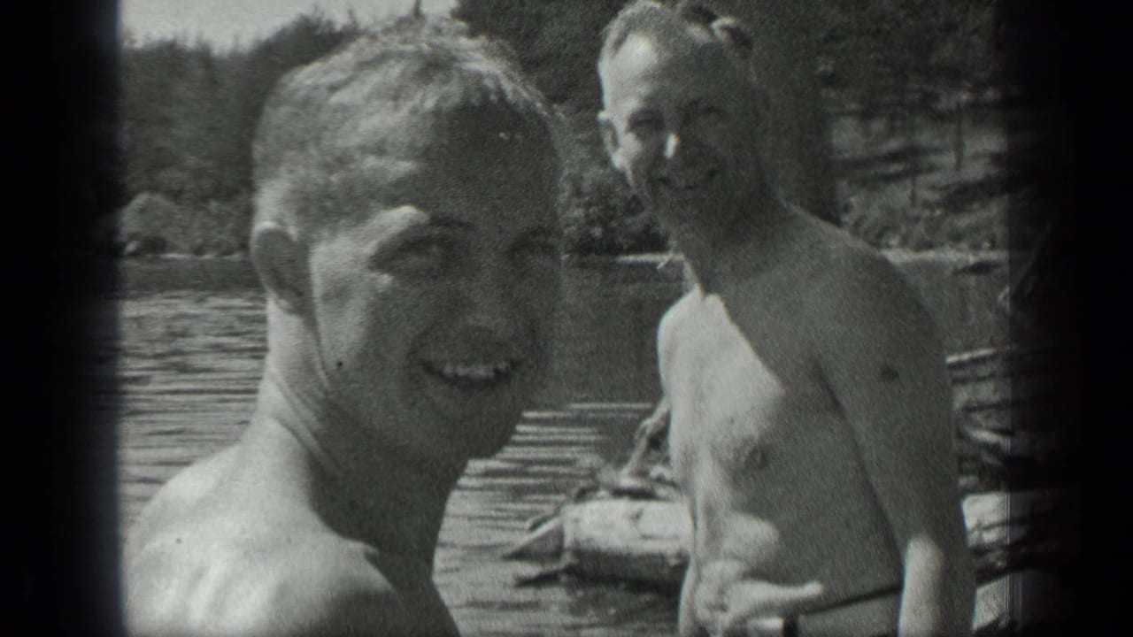 Two shirtless adult men stand or sit near a wooden dock in lake water.