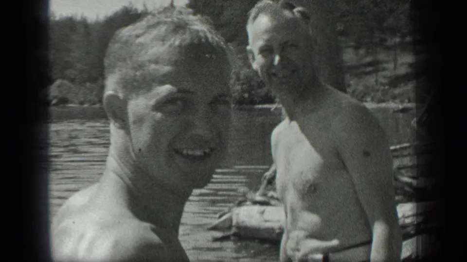 Two adult men in swimwear are seen by a wooden dock at Long Lake, Canada, with trees and rippled water behind them.