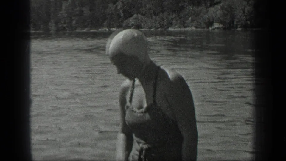 A woman in vintage swimwear and a cap stands at the water's edge at Long Lake, Canada, adjusting her cap in 1939 home-movie footage.