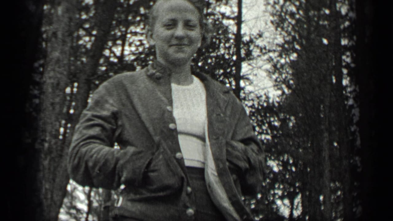 Woman smiling in a wooded area, wearing a jacket over a light top.
