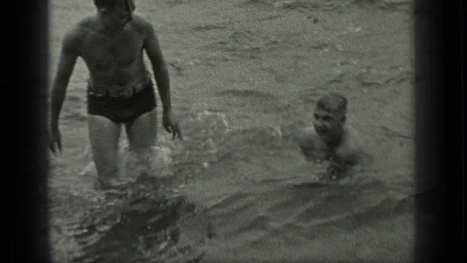 At Long Lake, Canada, one man in dark swim trunks stands in the water as another man swims beside him.