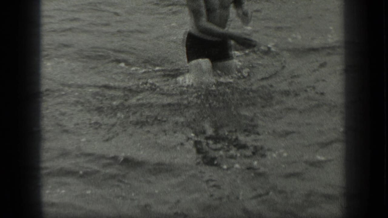 Man in dark swim trunks wading through shallow lake water.