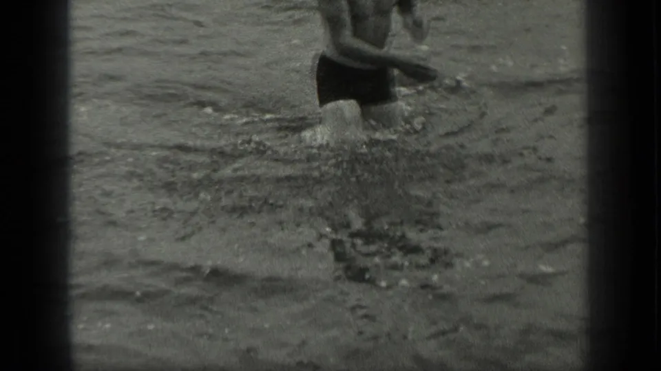 In 1939 Long Lake, Canada footage, a man in dark swim trunks wades through rippled shallow water near shore.