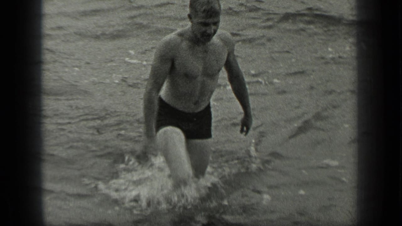 Shirtless man wading toward shore through splashing lake water.