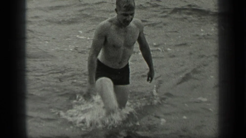 A shirtless adult man in swim trunks wades toward shore through splashing water at Long Lake, Canada, supported by clip metadata.