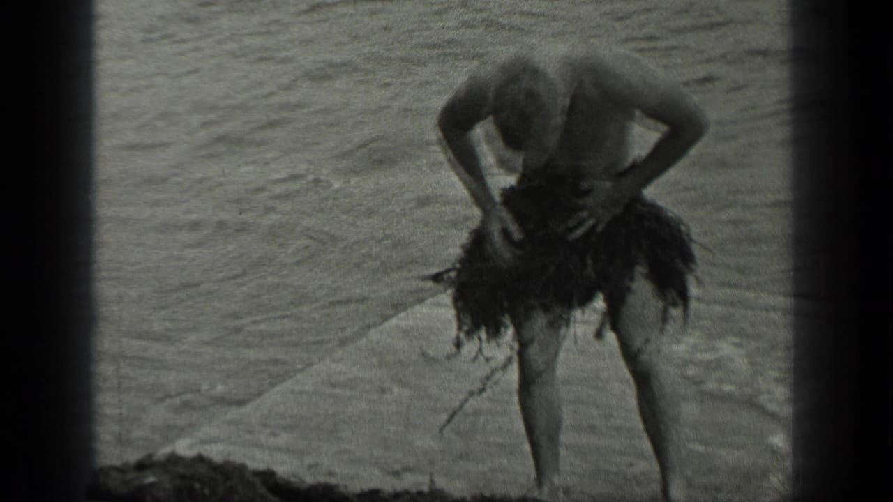 Shirtless man adjusts a seaweed skirt while standing near lake water.