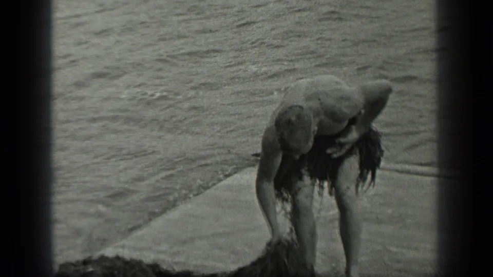 The man bends down beside the lake, holding or gathering seaweed for the improvised skirt in the 1939 Long Lake footage.