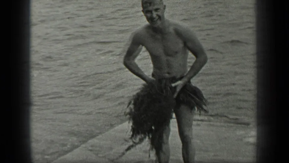 A shirtless man smiles toward the camera with seaweed arranged like a skirt on the Long Lake shoreline.