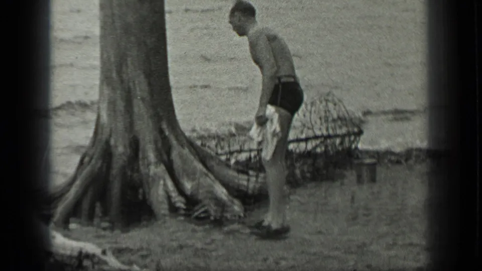 Near the lakeside, the man stands by a large tree trunk with a wire basket behind him and cloth or seaweed in his hands.