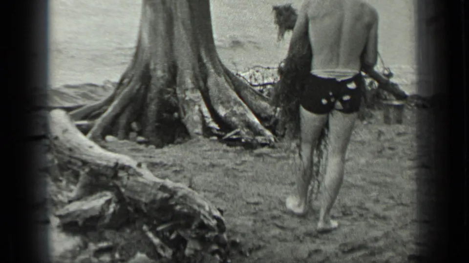 At Long Lake, Canada in 1939, a shirtless man in dark swim trunks walks barefoot past exposed roots while carrying branches.