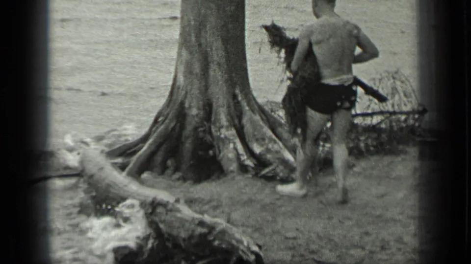 The man continues along the Long Lake shoreline, carrying a rough bundle near a large tree with exposed roots.
