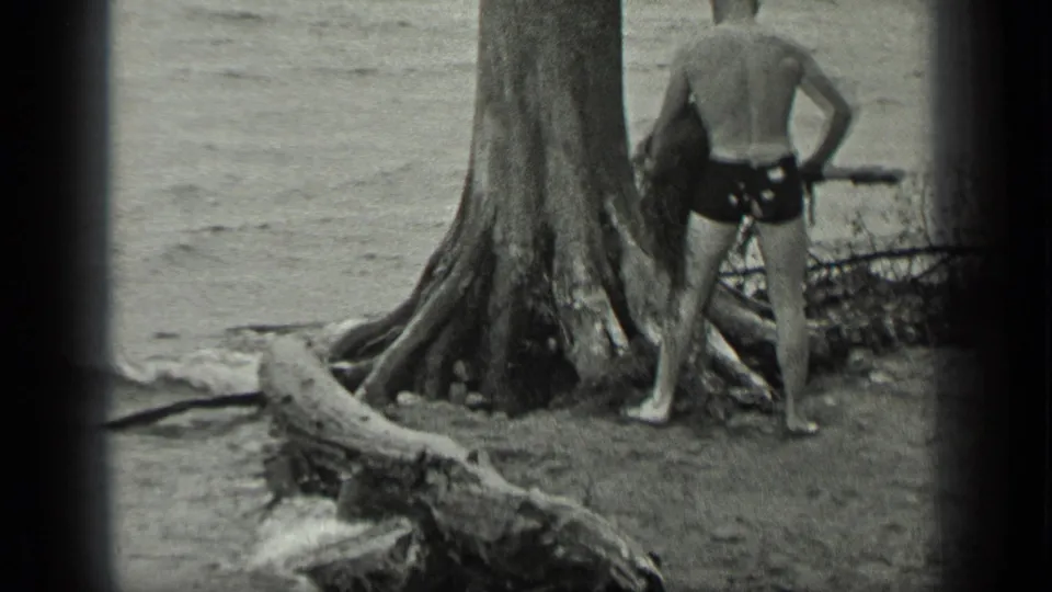 Near the Long Lake waterline, the man pauses by exposed tree roots while holding branches or root material.