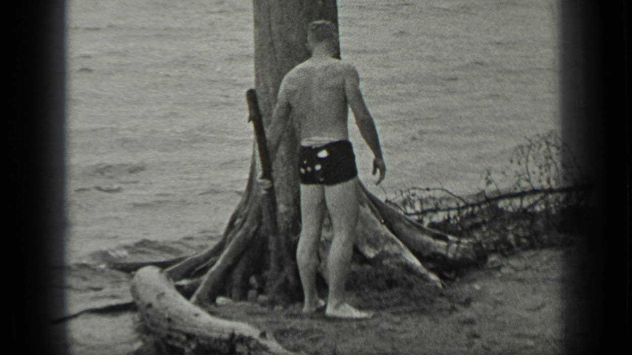 Shirtless man in swim trunks stands near a large lakeside tree with water behind him.