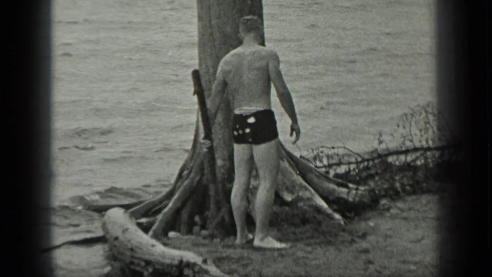 At Long Lake, Canada in 1939, a barefoot man in dark swim trunks stands beside a large shoreline tree near gentle water.