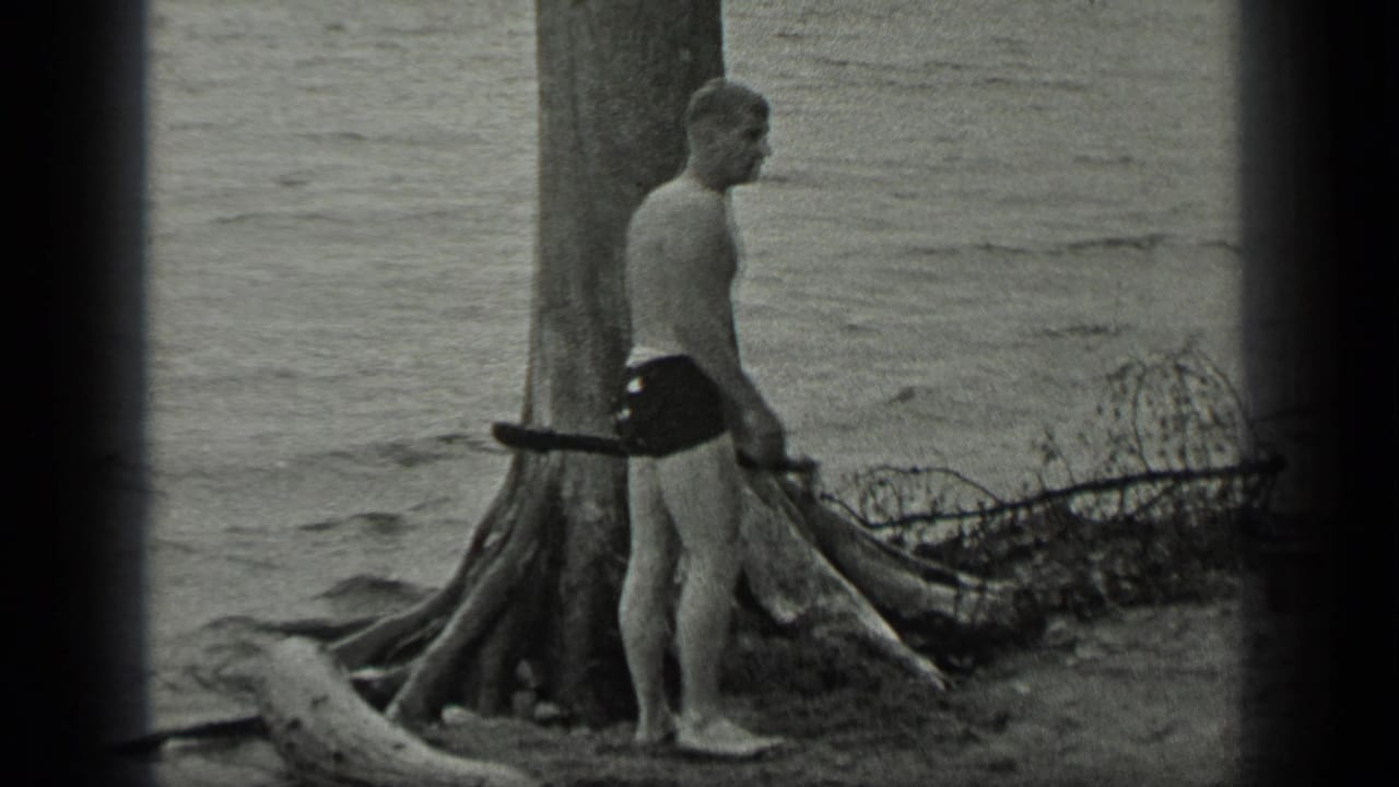 A barefoot man at Long Lake, Canada stands in profile beside a large tree, with a stick or branch near his side and water behind him.