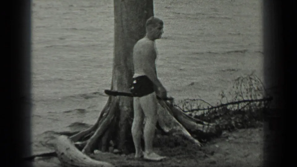 A barefoot man at Long Lake, Canada stands in profile beside a large tree, with a stick or branch near his side and water behind him.