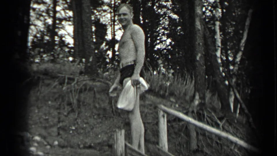 At Long Lake in 1939, a shirtless adult man in swim trunks holds a towel beside trees and a rough wooden railing.