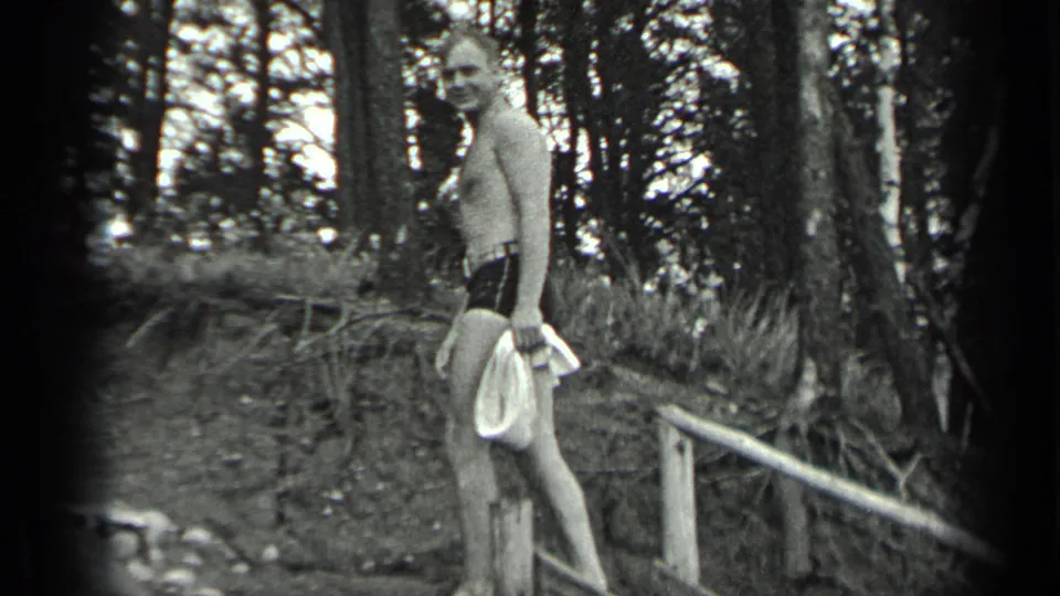 Black-and-white 1939 Long Lake frame of a man in swim trunks holding a towel on a wooded lakeside access path.