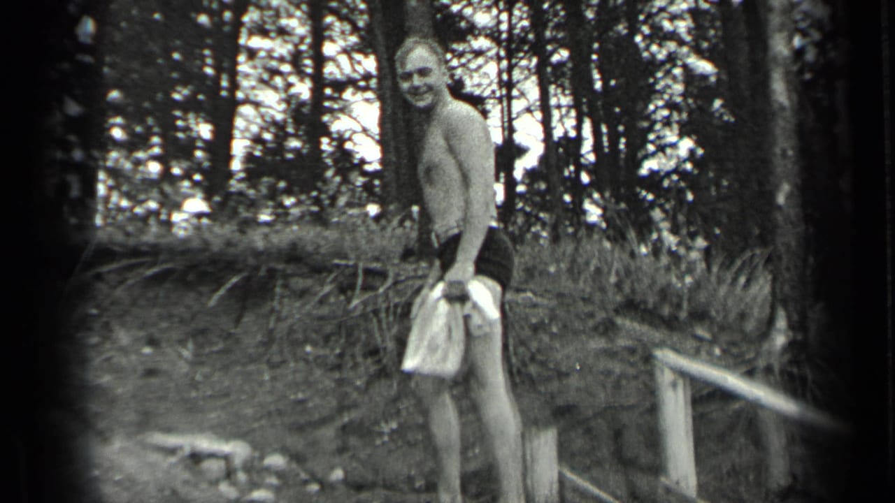Shirtless man in swim trunks holding a towel on a wooded lakeside bank.