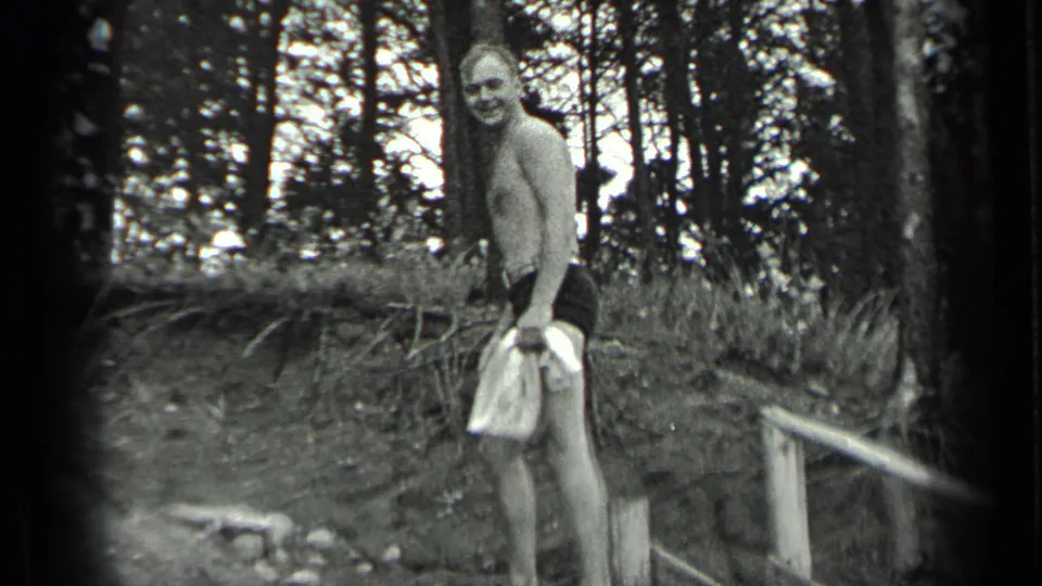 In 1939 Long Lake home-movie footage, a shirtless man in swim trunks smiles while holding a towel near trees and a rough lakeside bank.