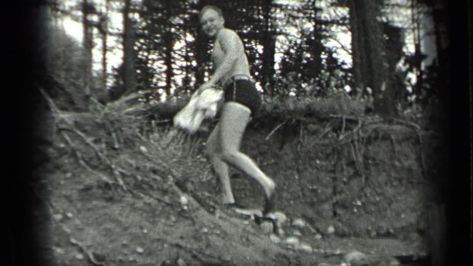 The man descends a sloped wooded bank near Long Lake, holding a towel and looking toward the camera in black-and-white 8mm footage.