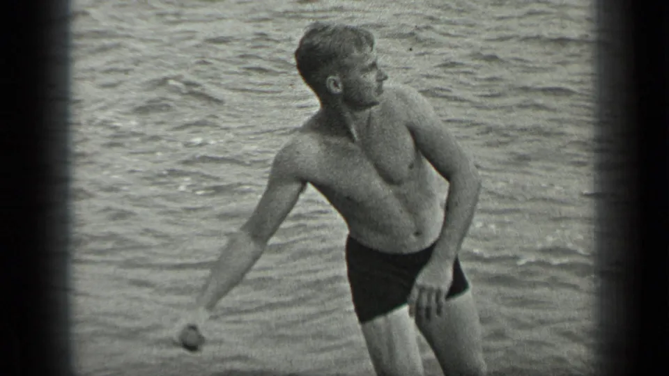 A shirtless man in swim trunks stands in shallow lake water and winds up with a small stone, with rippled water behind him.