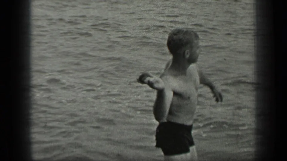 The man follows through after skipping a rock across the lake surface, framed by rippled water and dark 8mm film edges.