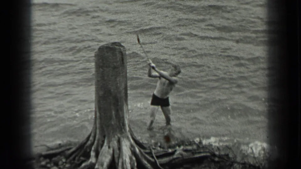 At Long Lake, Canada in 1939, a shirtless man raises an axe near driftwood, a large stump, and rippled water.