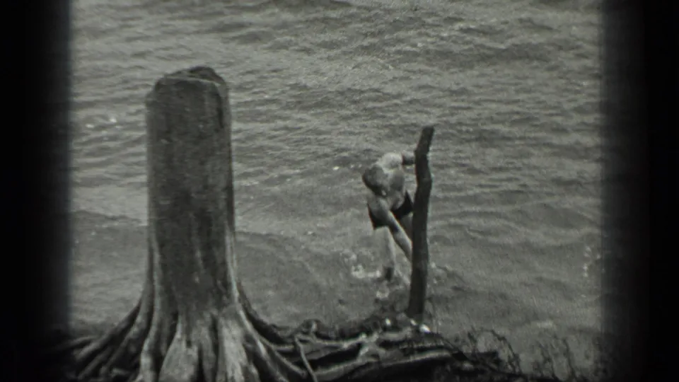 In 1939 Long Lake, Canada footage, a man in swim trunks reaches toward a log in shallow water beside a large shoreline stump.