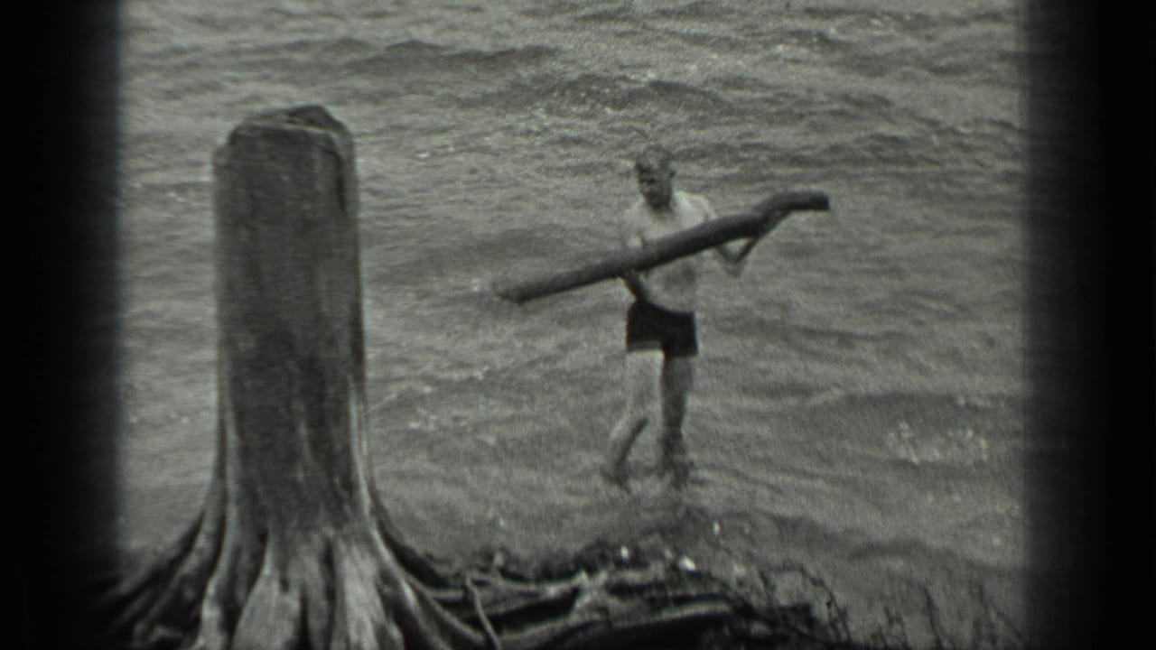 Man wades through shallow water carrying a long log across his shoulders.