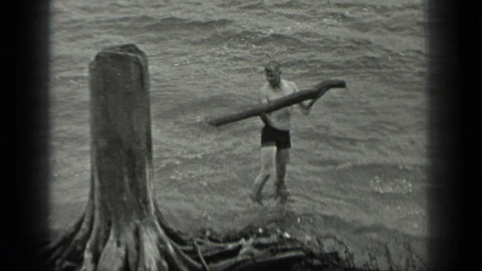 A man wades in Long Lake water with a long log balanced across his shoulders, with a tree stump and shoreline in view.