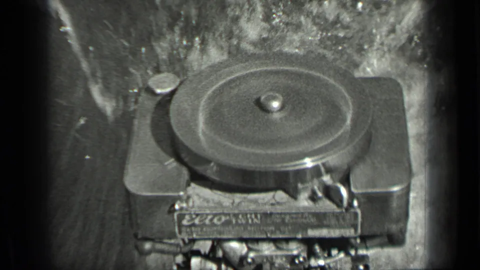 Close-up of a film projector or reel mechanism in black-and-white 8mm footage, with a partial marking visible on the machine.