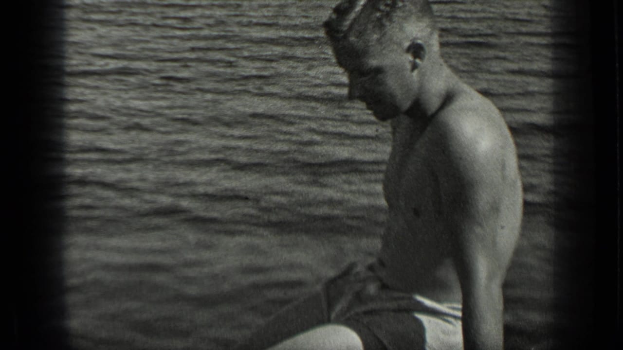 Shirtless man in swim trunks seated in a small boat on the lake.