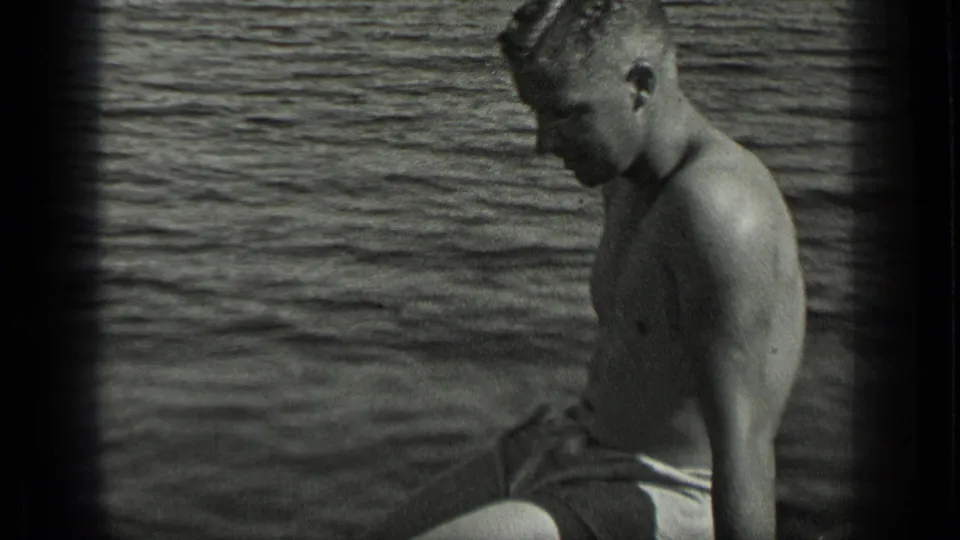 A man in swim trunks sits in a small boat on Long Lake, Canada, looking downward against rippled water.