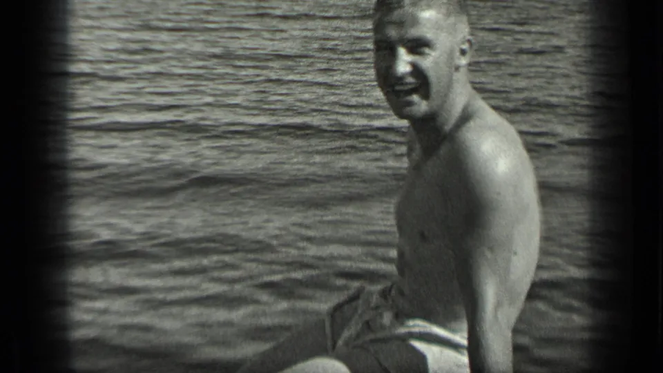 The seated man turns toward the camera in a black-and-white Long Lake home-movie frame, with rippled water behind him.