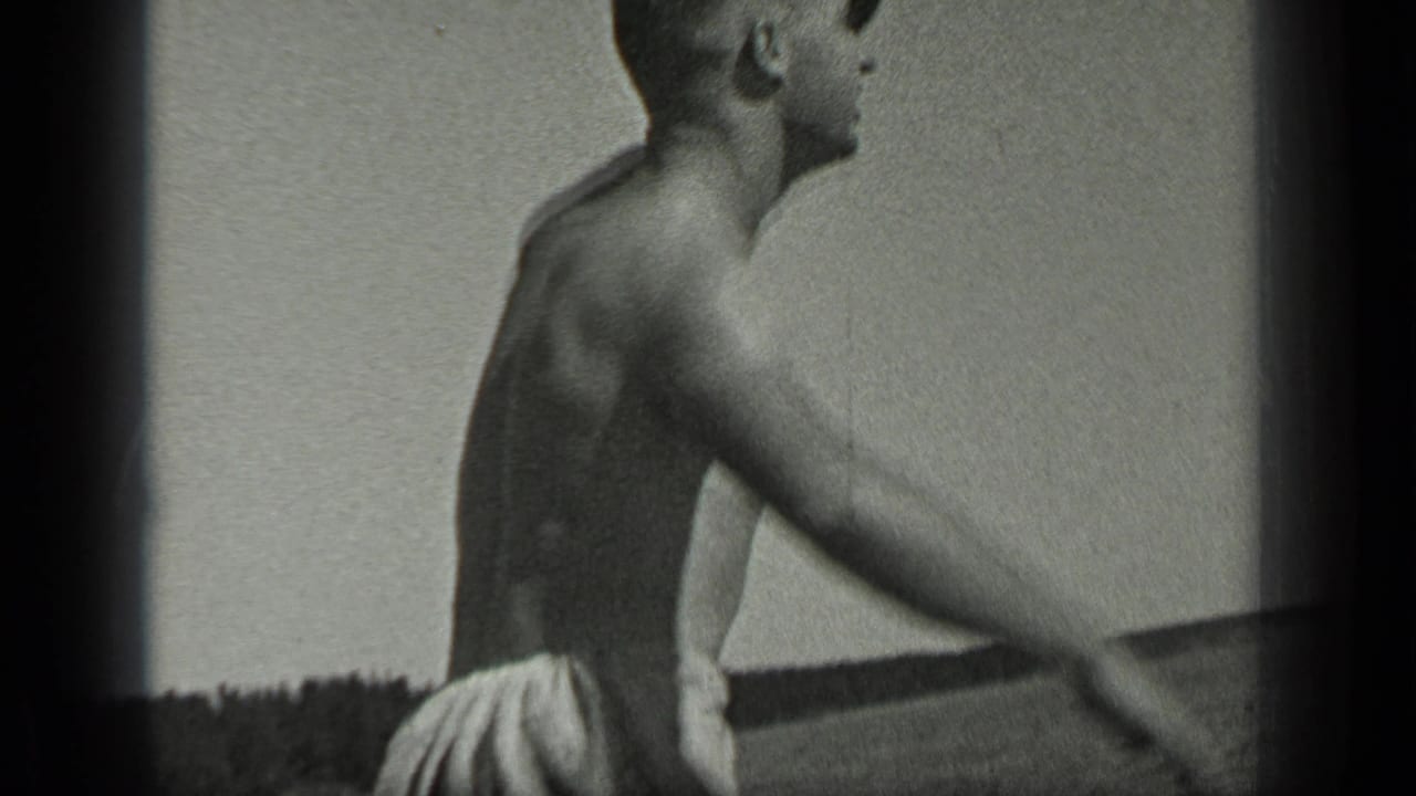 Shirtless man in swim trunks seen from the side beside open lake water.
