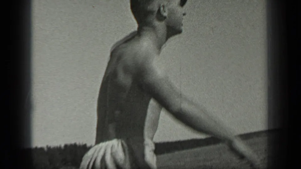 Side view from 1939 Long Lake footage showing a shirtless man in swim trunks near the water with distant shoreline behind him.