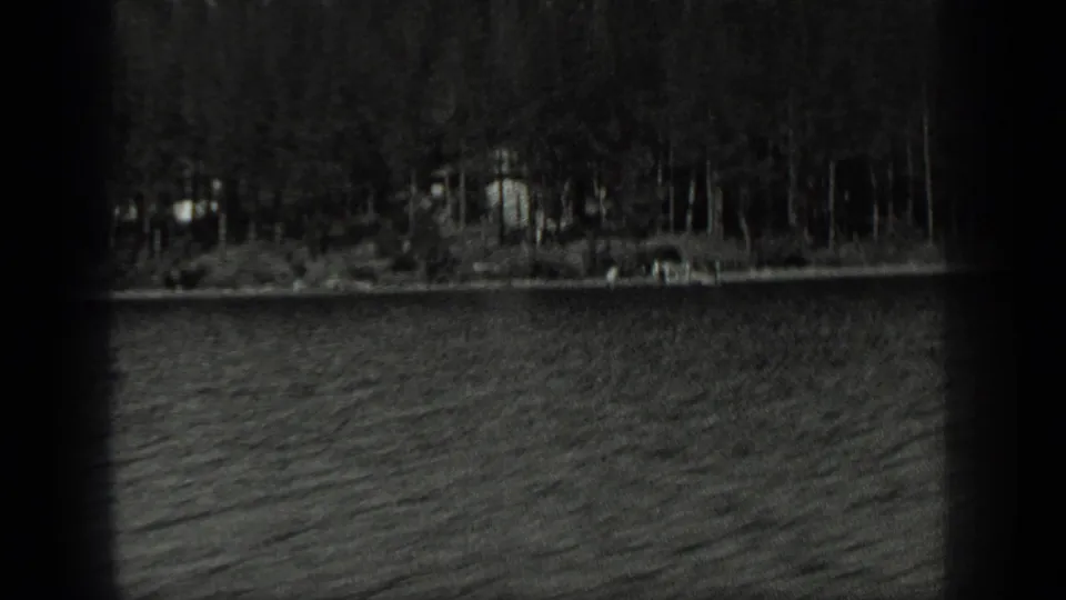 Grainy 8mm view across Long Lake, Canada, with rippled water, dense trees and small distant lakeside structures on shore.