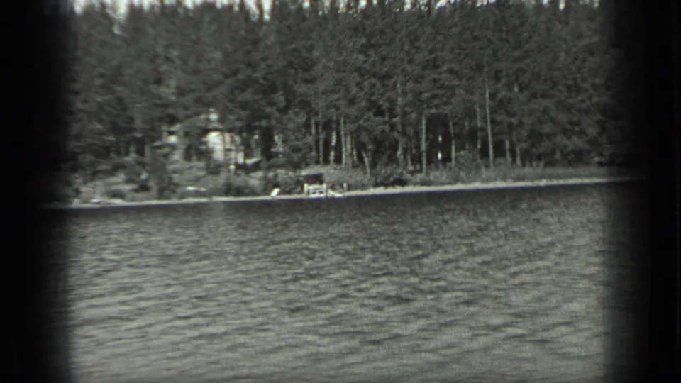 Wide 1939 Long Lake view showing rippled water, dense forest, a small shoreline structure and home-movie frame edges.