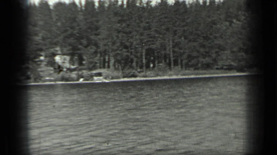 1939 Long Lake, Canada home-movie frame showing rippled water, dense trees, and a small pale lakeside structure on the shore.