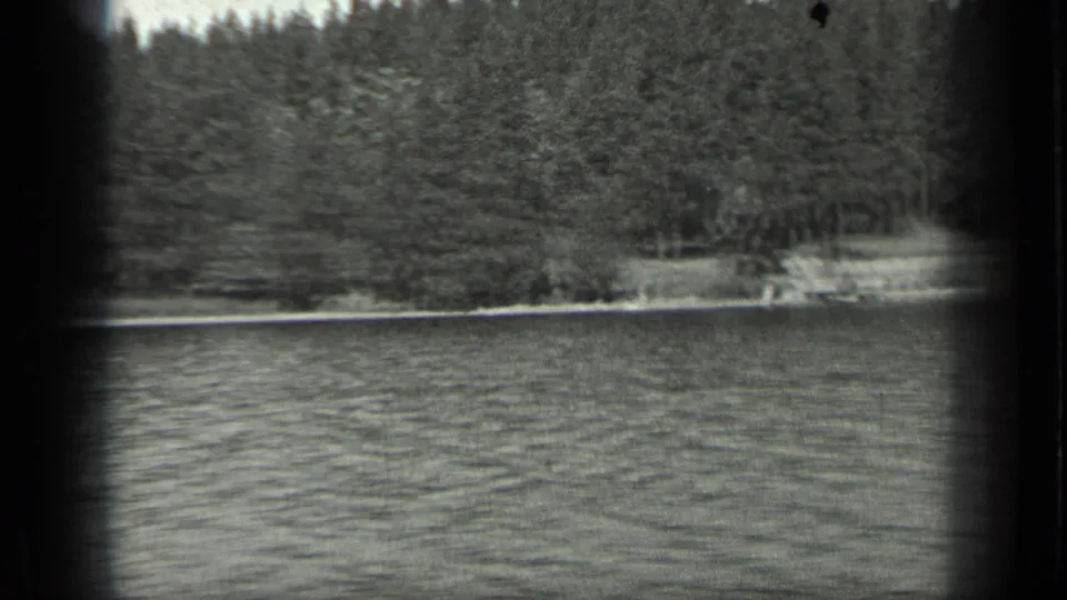Black-and-white 8mm view of Long Lake, Canada, with rippled water and a shadowed forest bank passing across the frame.