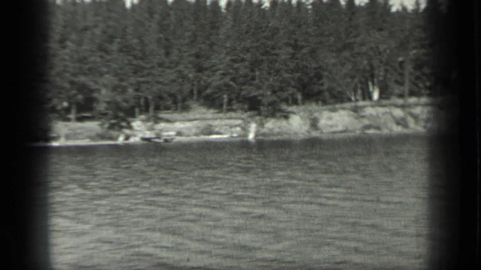 1939 Long Lake frame showing a forested shore, pale bank, and small indistinct lakeside objects beyond the rippled water.