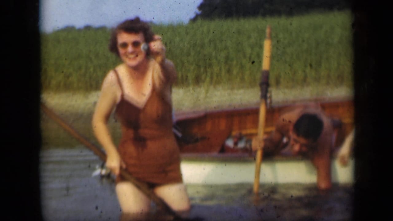 Woman in a brown swimsuit smiles in shallow water beside a wooden rowboat.