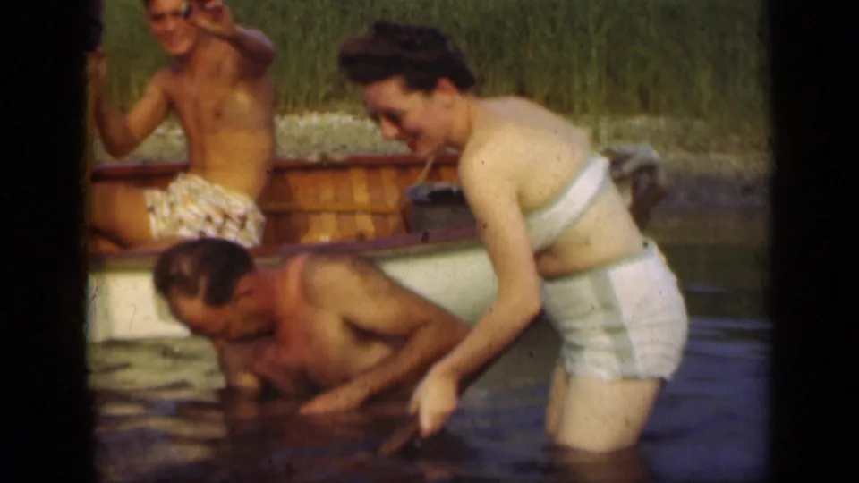 A woman in a white swimsuit bends toward the water as a man leans low beside a wooden rowboat in 1948 Long Island home-movie footage.