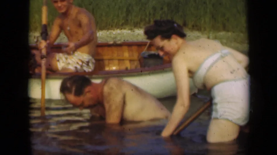 Adults in swimsuits work or play in shallow water beside a wooden rowboat, with a woman using a long handle near the surface.