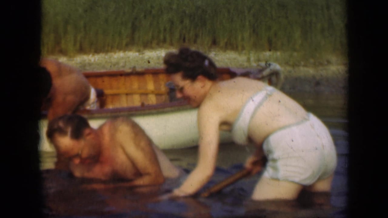 1948 Long Island home-movie frame of a woman standing by a crouched man beside a rowboat in shallow lake water.