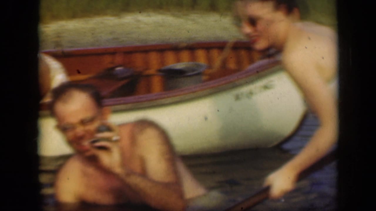 Shirtless man with glasses holds a small object near a canoe as a woman stands nearby.